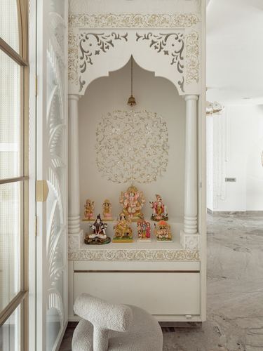 Carved white arched pooja niche with deity idols and hanging brass bell, boucle stool in front