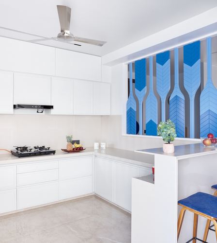White modular kitchen with L-shaped counter, gas stove, and blue chevron-patterned glass window above bar counter