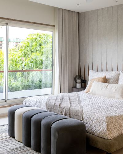 Bedroom with fluted wood headboard wall, layered white bedding, and rounded grey velvet bench facing large window