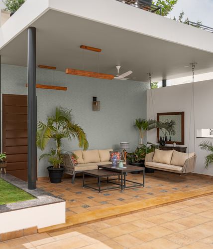 Covered patio seating area with hanging swing, sofa, and potted palms against a textured grey wall