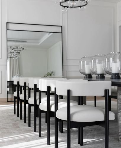 Row of black-legged white dining chairs beside a large leaning mirror and glass candle holders