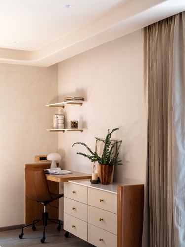 Corner study nook with wooden desk, leather chair, floating shelves and drawer unit against beige wall