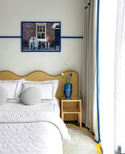 Bedroom corner with mustard curved headboard, white embroidered bedding, framed photo art and cane side table