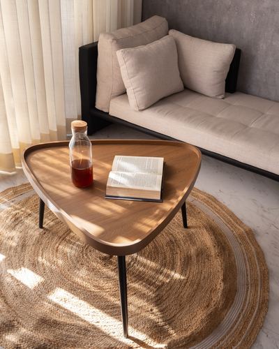 Triangular wood coffee table with book and bottle on jute rug beside beige sofa