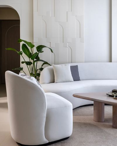 Curved white sofa and armchair beside round stone coffee table, fluted plaster wall behind
