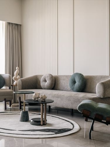 Beige sofa with round bolster cushions beside stacked round tables on a circular rug, tall paneled wall behind
