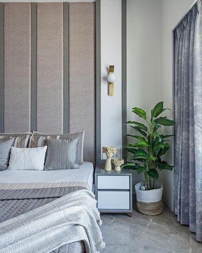 Bedroom corner with channeled upholstered headboard, brass sconce, bedside table, and potted plant near a curtained window
