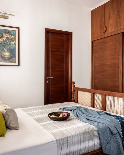 Bedroom corner with cane headboard, teak door, wardrobe, and blue throw on bed