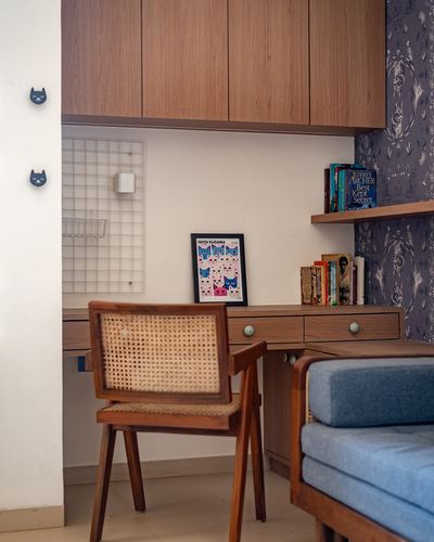 Wooden study nook with cane chair, floating shelf of books, and a Yayoi Kusama cat poster against dark floral wallpaper