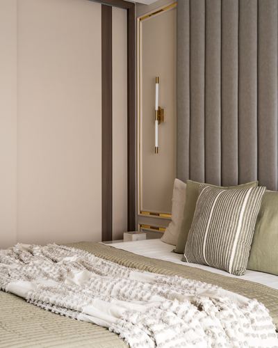Bedroom corner with channel-tufted taupe headboard, olive striped cushions and a brass wall sconce beside a paneled wardrobe.