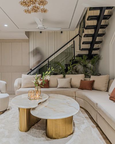 Curved beige sectional and brass-based marble coffee tables beneath a floating staircase with planting below