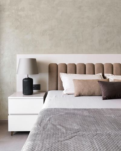 Bed with channel-tufted taupe headboard, white nightstand, dark ribbed lamp, textured plaster wall behind.