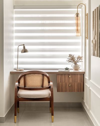Cane-back wooden chair at a floating walnut desk before a sunlit striped blind, with brass sconce and lamp.