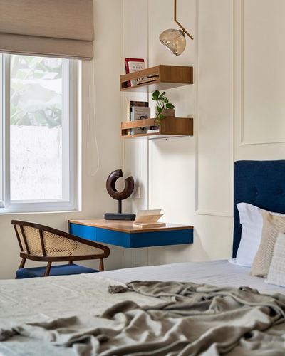 Bedroom corner with navy headboard, floating blue nightstand, wooden shelves and cane chair by window