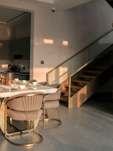 Dining table with gold wire chairs beside an open wooden staircase, warm evening light on plaster wall