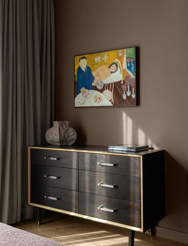 Dark wood dresser with brass trim beneath a colorful figurative painting in a warm-toned bedroom corner