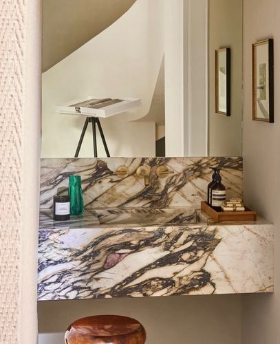 Floating veined marble vanity with brass faucet, mirror reflecting a curved staircase and easel artwork.