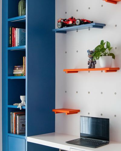 Study nook with navy bookshelf, orange floating shelves, plant, toy car, and laptop on white desk