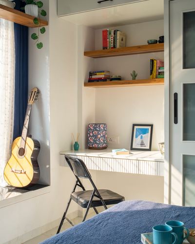 Sunlit study nook with floating desk, wall shelves, folding chair, and guitar by window