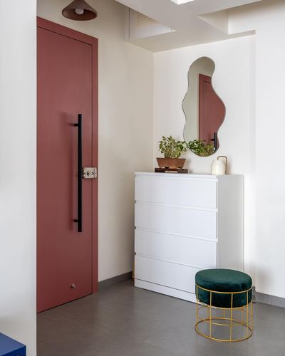 Entryway with terracotta door, white dresser, wavy mirror, and green velvet stool on brass legs