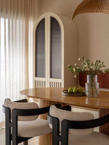Oval wood dining table with boucle chairs beneath an arched fluted cabinet