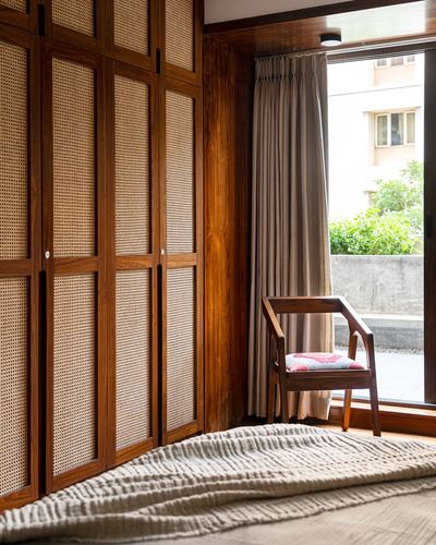 Bedroom corner with cane wardrobe doors, wooden armchair, and window looking onto terrace greenery