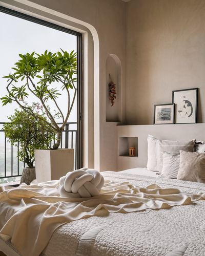 Minimalist beige bedroom with arched niche, layered bedding, and balcony view of a frangipani tree