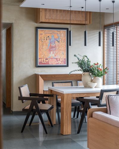 Dining area with wooden table, cane chairs, pendant lights, and framed Indian folk artwork on plastered wall