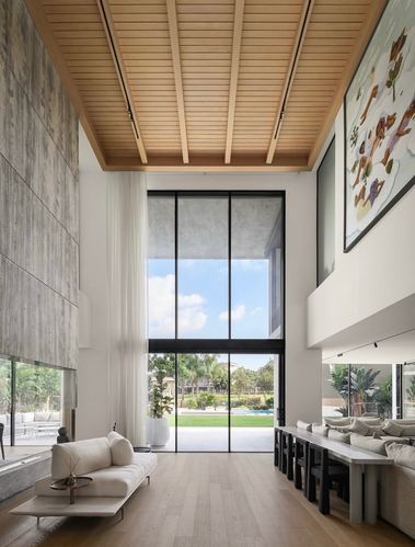 Double-height living room with wood-slat ceiling, floor-to-ceiling windows overlooking a garden and pool, beige sofa in foreground