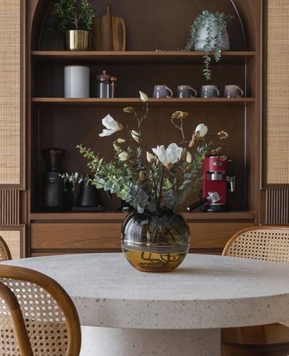 Round terrazzo dining table with cane chairs in front of an arched wooden shelving unit holding mugs and an espresso machine