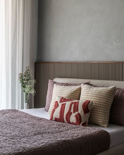 Bedroom corner with fluted wood headboard, mauve quilted bedspread, and textured cushions beside sheer curtain