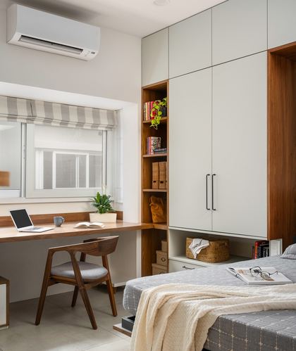 Bedroom corner with built-in study desk under window and tall wardrobe with open wood shelving beside bed.