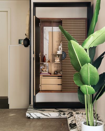 Black-framed pooja unit with textured metal door, deity figurines, and marble plinth beside a large plant