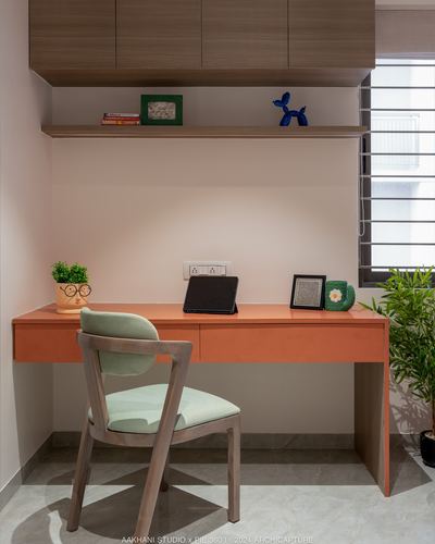 Study nook with orange desk, wooden chair, and floating shelf under overhead cabinets near window