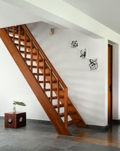 Wooden staircase rising diagonally against white wall with three abstract artworks in stone-floored hallway