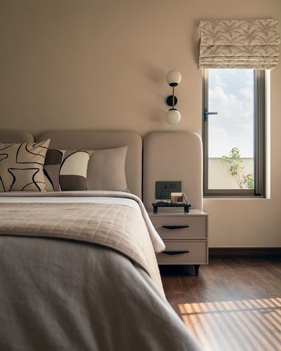 Beige bedroom corner with upholstered headboard, bedside table, sconce, and sunlit window