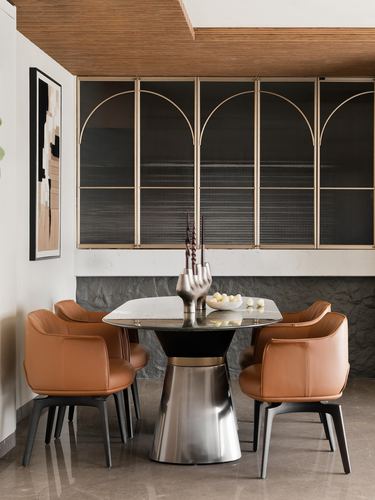 Round marble dining table with tan leather chairs beneath an arched glass and brass cabinet