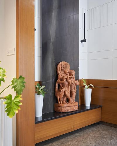 Terracotta figurine sculpture on a black stone bench set within a dark stone wall niche flanked by wood panelling and plants