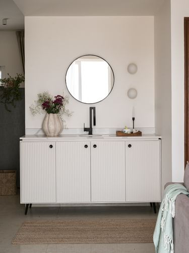 White fluted vanity cabinet with round mirror, black faucet, and vase of flowers in a niche wall