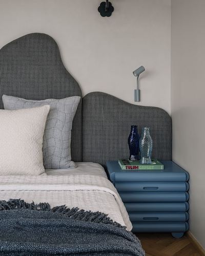 Bed with scalloped grey headboard beside a ribbed blue nightstand and reading lamp