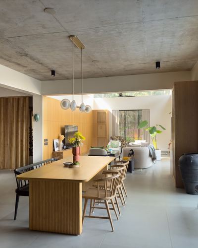 Open-plan dining and living space with wooden table, cane chairs, exposed concrete ceiling, and view to greenery beyond.