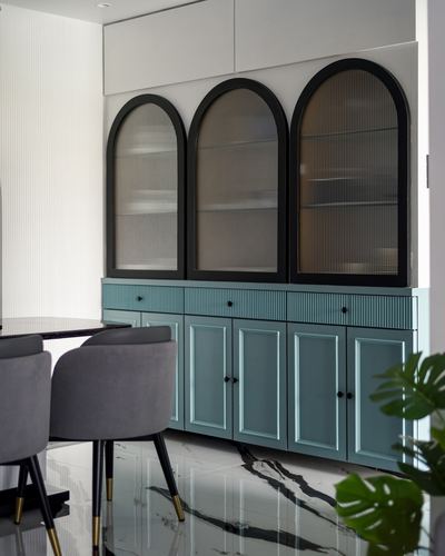 Teal cabinet with three arched black-framed glass doors above fluted drawers, dining chairs in foreground