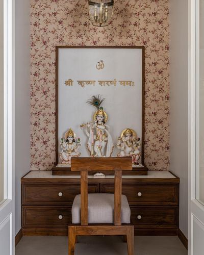 Small pooja alcove with floral wallpaper, marble idol backdrop, wooden dresser and chair below