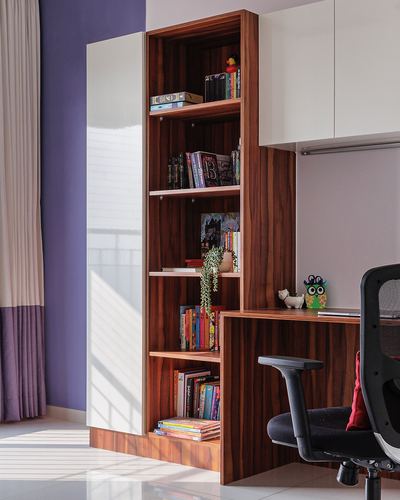Wood-veneer bookshelf built into a study desk beside a lavender wall and mesh office chair