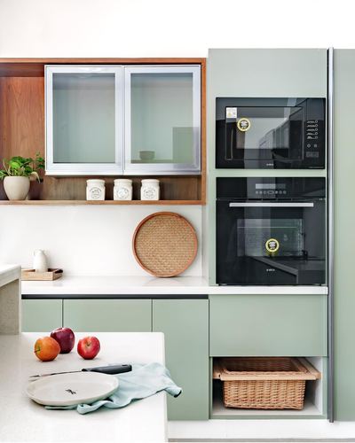 Sage green kitchen cabinetry with built-in oven, microwave, wood open shelf and wicker basket storage