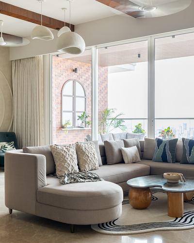 Curved beige sectional sofa with round wood-and-glass tables in a sunlit living room with brick accent wall