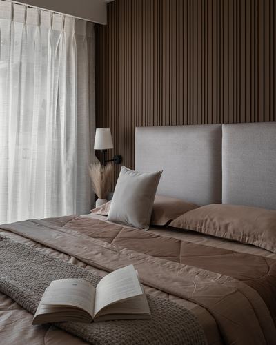Bedroom with fluted wood wall, grey upholstered headboard, brown linens, sheer curtains and an open book