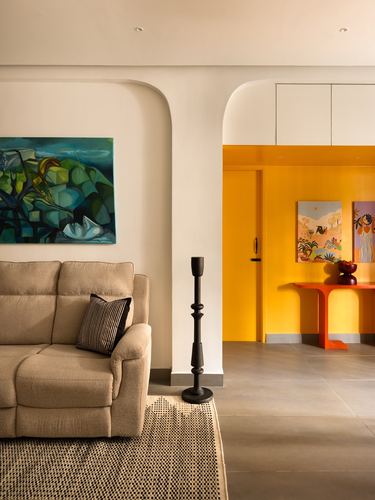Living room with beige sofa and green painting opens through arch to yellow hallway with art and orange console