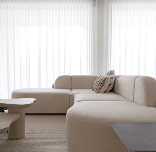 Cream boucle sectional sofa with patterned pillow near sheer curtained windows and sculptural stone side table