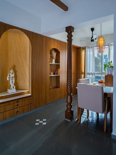 Wood-panelled pooja niche with deity statue beside carved column and dining area